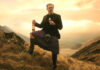 A man in a kilt stands against a sunset in the Highlands with a bottle of Ben Bracken whisky in his hand and a glass with the whisky inside in the other.