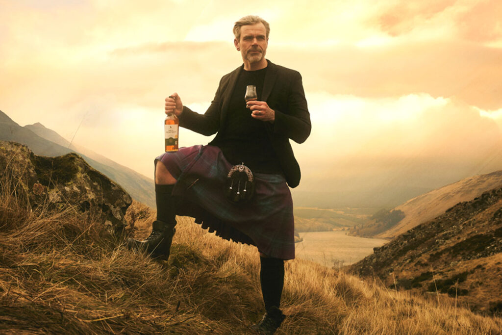 A man in a kilt stands against a sunset in the Highlands with a bottle of Ben Bracken whisky in his hand and a glass with the whisky inside in the other.