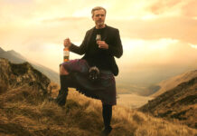 A man in a kilt stands against a sunset in the Highlands with a bottle of Ben Bracken whisky in his hand and a glass with the whisky inside in the other.