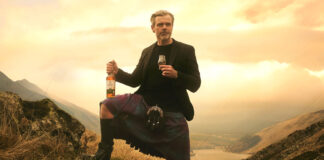 A man in a kilt stands against a sunset in the Highlands with a bottle of Ben Bracken whisky in his hand and a glass with the whisky inside in the other.