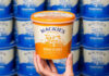 A hand holds up a tub of Mackie's of Scotland Honeycomb Ice Cream with tubs of Mackie's of Scotland Traditional lining the wall in the background.