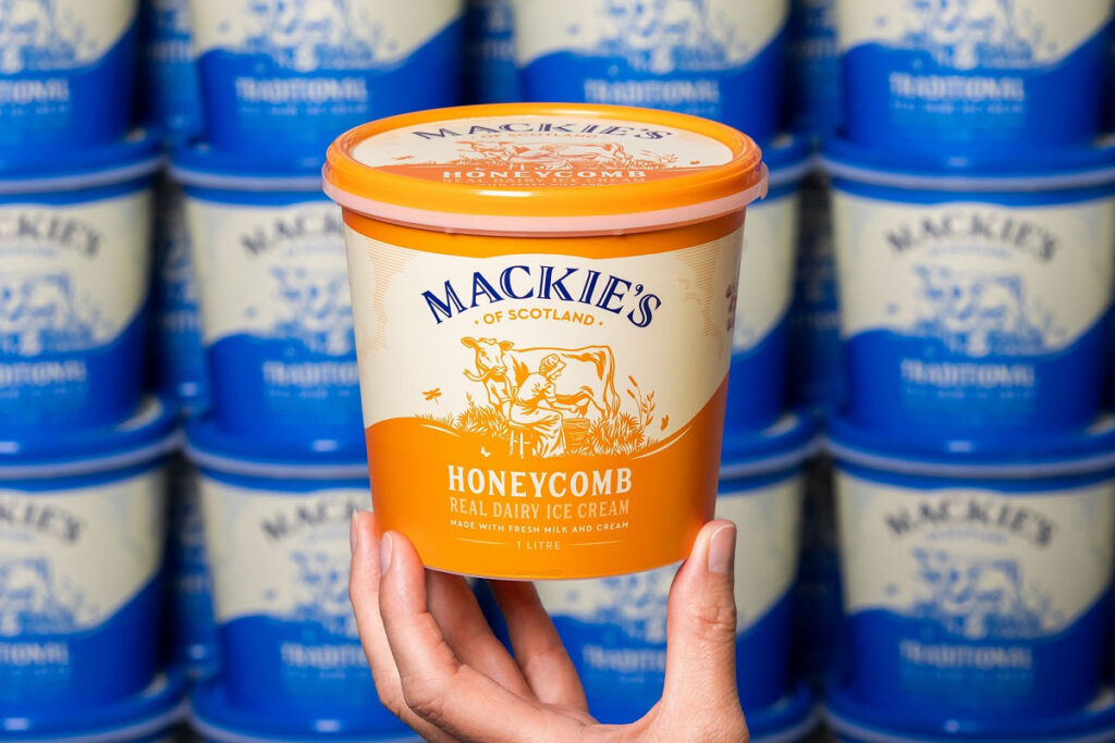 A hand holds up a tub of Mackie's of Scotland Honeycomb Ice Cream with tubs of Mackie's of Scotland Vanilla lining the wall in the background.