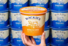 A hand holds up a tub of Mackie's of Scotland Honeycomb Ice Cream with tubs of Mackie's of Scotland Traditional lining the wall in the background.