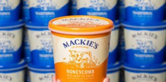 A hand holds up a tub of Mackie's of Scotland Honeycomb Ice Cream with tubs of Mackie's of Scotland Traditional lining the wall in the background.