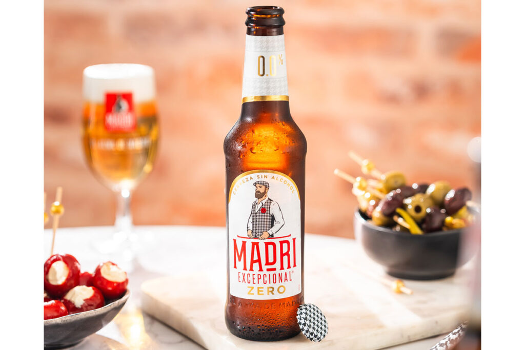 A bottle of Madrí Excepcional Zero stands on a table with a bowl of olives to the right and a bowl of cheese stuffed tomatoes to the left with a Madrí Excepcional branded glass behind the bottle.