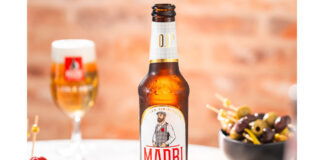 A bottle of Madrí Excepcional Zero stands on a table with a bowl of olives to the right and a bowl of cheese stuffed tomatoes to the left with a Madrí Excepcional branded glass behind the bottle.