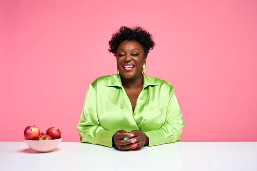 British comedian Judi Love sits at a white table wearing a bright green top with bowl of Pink Lady apples to her right.