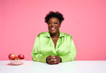 British comedian Judi Love sits at a white table wearing a bright green top with bowl of Pink Lady apples to her right.