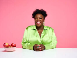 Pink Lady teams up with Judi Love to empower the lunch hour British comedian Judi Love sits at a white table wearing a bright green top with bowl of Pink Lady apples to her right.