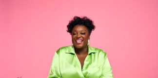 British comedian Judi Love sits at a white table wearing a bright green top with bowl of Pink Lady apples to her right.