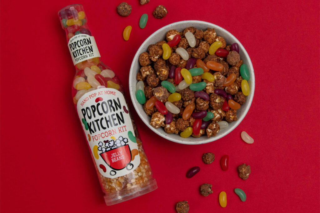 A bottle of Popcorn Kitchen Jelly Beans lies against a red background with a bowl of chocolate covered popcorn and jelly beans to the right of it, with some of the popcorn and sweets spilling out and onto the red background.