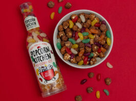 A bottle of Popcorn Kitchen Jelly Beans lies against a red background with a bowl of chocolate covered popcorn and jelly beans to the right of it, with some of the popcorn and sweets spilling out and onto the red background.