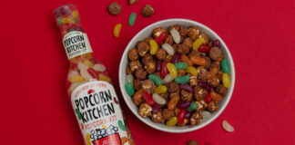 A bottle of Popcorn Kitchen Jelly Beans lies against a red background with a bowl of chocolate covered popcorn and jelly beans to the right of it, with some of the popcorn and sweets spilling out and onto the red background.