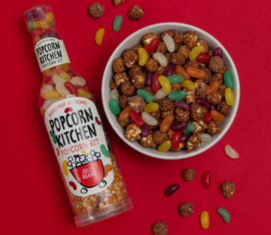 Popcorn Kitchen brings a multi-texture snacks treat A bottle of Popcorn Kitchen Jelly Beans lies against a red background with a bowl of chocolate covered popcorn and jelly beans to the right of it, with some of the popcorn and sweets spilling out and onto the red background.