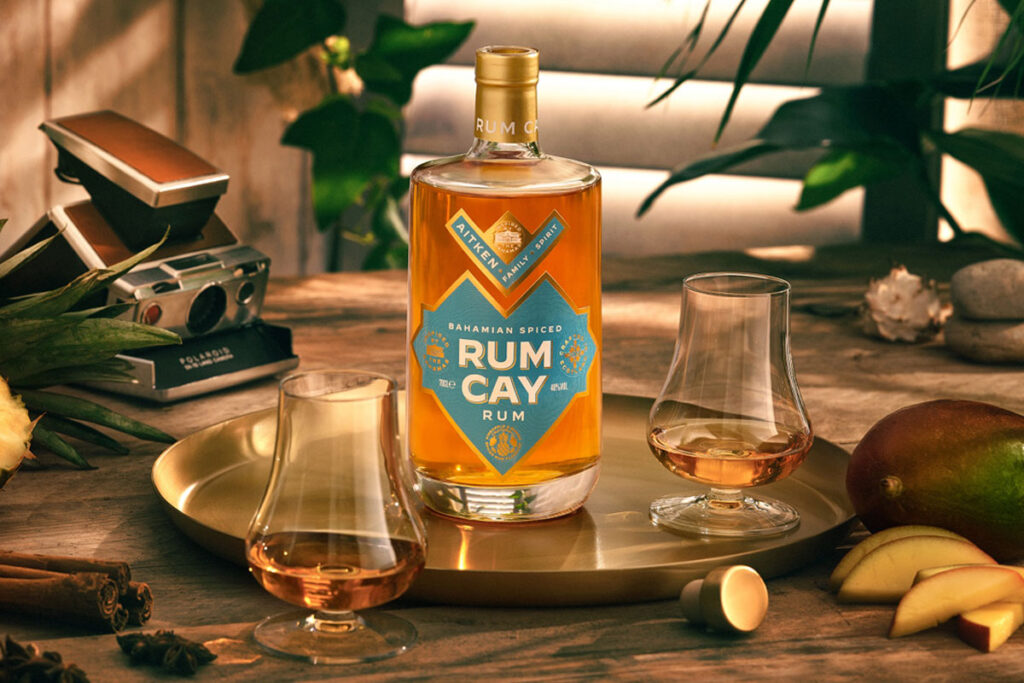 A bottle of Rum Cay Rum is standing on a serving plate on a table with the spirit poured out into nosing glasses, the tray has mangoes and a cut open pineapple around it there is a camera set behind the tray.