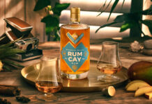 A bottle of Rum Cay Rum is standing on a serving plate on a table with the spirit poured out into nosing glasses, the tray has mangoes and a cut open pineapple around it there is a camera set behind the tray.