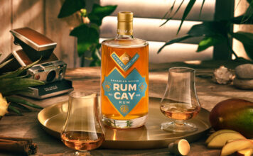 A bottle of Rum Cay Rum is standing on a serving plate on a table with the spirit poured out into nosing glasses, the tray has mangoes and a cut open pineapple around it there is a camera set behind the tray.