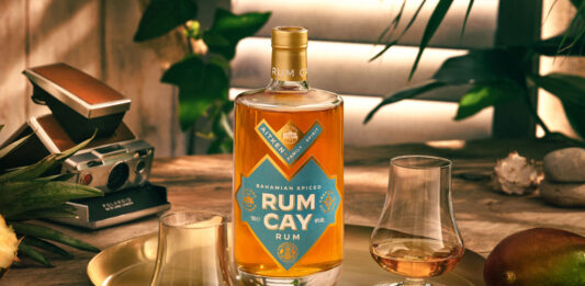 A bottle of Rum Cay Rum is standing on a serving plate on a table with the spirit poured out into nosing glasses, the tray has mangoes and a cut open pineapple around it there is a camera set behind the tray.