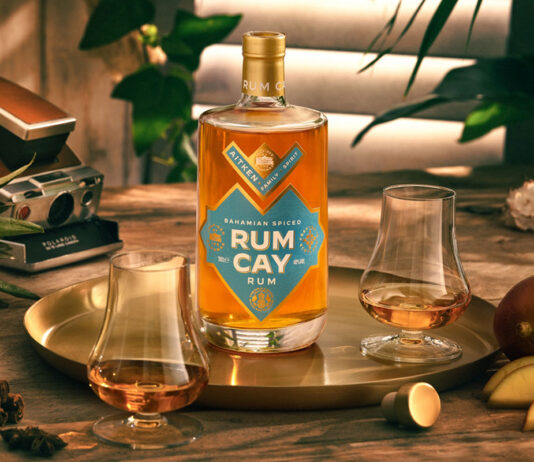 Scottish Bahamian spiced Rum Cay Rum launches A bottle of Rum Cay Rum is standing on a serving plate on a table with the spirit poured out into nosing glasses, the tray has mangoes and a cut open pineapple around it there is a camera set behind the tray.