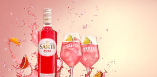 A bottle of Sarti Rosa splashes down in a liquid with two wine glasses next to it fill with the spirit and fruit, with pieces of fruit splashing into the liquid as well.