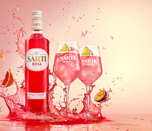 Sarti Rosa aperitif lands in the UK A bottle of Sarti Rosa splashes down in a liquid with two wine glasses next to it fill with the spirit and fruit, with pieces of fruit splashing into the liquid as well.