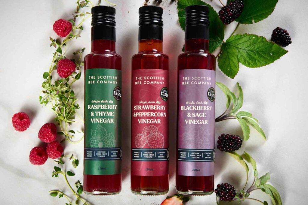 Bottles of The Scottish Bee Company Honey Drizzles lie across a marble countertop with raspberry plants and blackberry plants around the bottles.