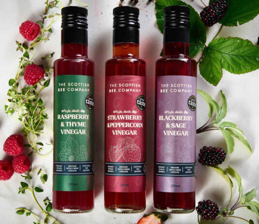 Scottish Bee Company unveils its new look for 2025 Bottles of The Scottish Bee Company Honey Drizzles lie across a marble countertop with raspberry plants and blackberry plants around the bottles.