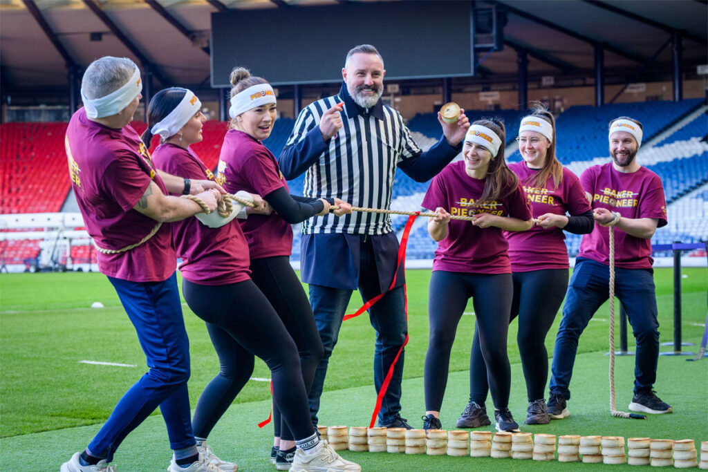 A group of people play Tug of War in Hampden stadium with a man in the middle of the group wearing a referee outfit and holding a Scotch pie with a small wall of Scotch pies in between the two groups.