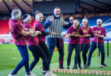 A group of people play Tug of War in Hampden stadium with a man in the middle of the group wearing a referee outfit and holding a Scotch pie with a small wall of Scotch pies in between the two groups.