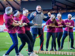 A group of people play Tug of War in Hampden stadium with a man in the middle of the group wearing a referee outfit and holding a Scotch pie with a small wall of Scotch pies in between the two groups.