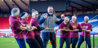 A group of people play Tug of War in Hampden stadium with a man in the middle of the group wearing a referee outfit and holding a Scotch pie with a small wall of Scotch pies in between the two groups.