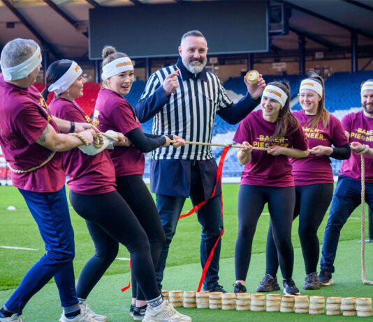 Final call to enter the World Championship Scotch Pie Awards A group of people play Tug of War in Hampden stadium with a man in the middle of the group wearing a referee outfit and holding a Scotch pie with a small wall of Scotch pies in between the two groups.