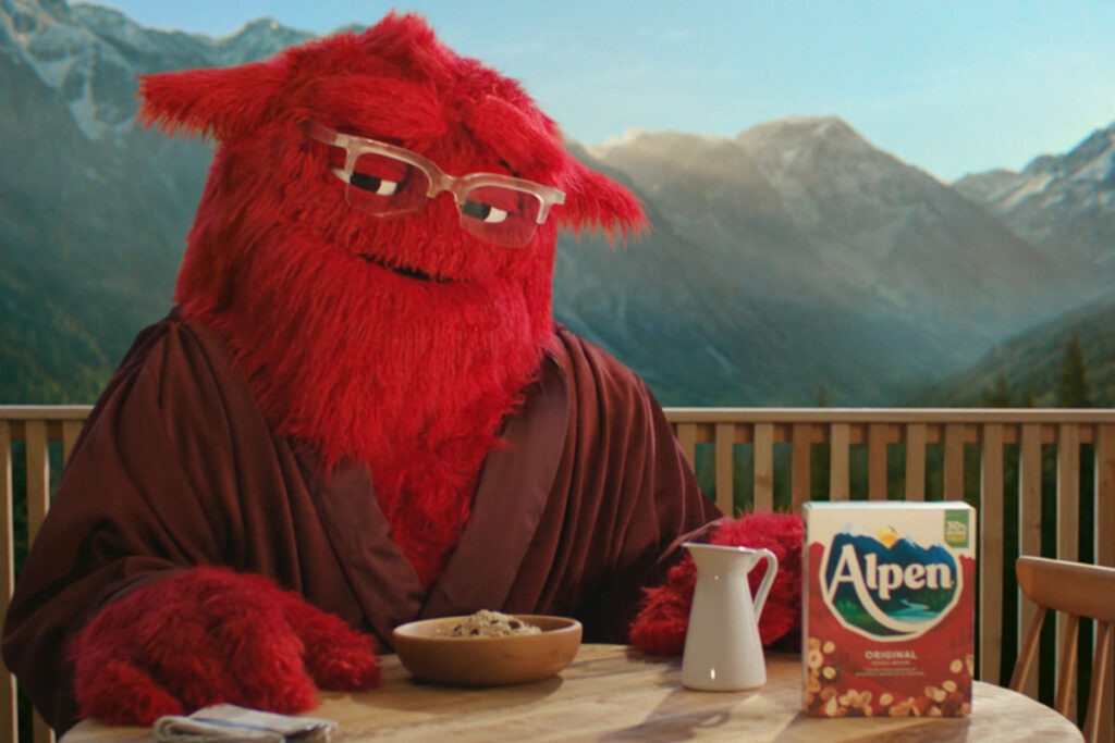 Alpen's mascot 'Ralph' sits at a breakfast table with a box of Alpen, a jug for milk and a bowl of muesli in front of her. Ralph is a large, red and furry character who wears glasses.