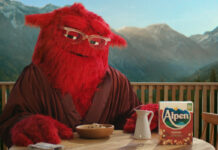 Alpen's mascot 'Mother' sits at a breakfast table with a box of Alpen, a jug for milk and a bowl of muesli in front of her. Mother is a large, red and furry character who wears glasses.