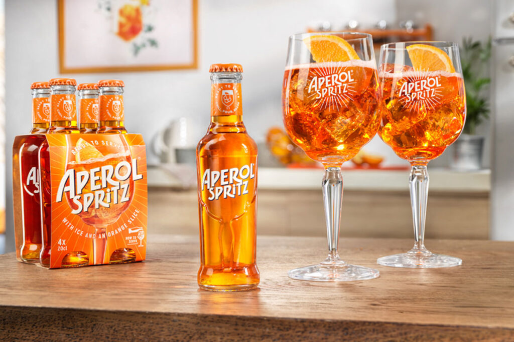 A bottle of Aperol Spritz RTD sits in the middle of a table, with two balloon glasses filled with the cocktail to the bottle's right and a four pack of bottles to the single bottle's left.