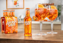 A bottle of Aperol Spritz RTD sits in the middle of a table, with two balloon glasses filled with the cocktail to the bottle's right and a four pack of bottles to the single bottle's left.