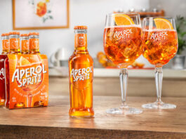 Aperol Spritz comes to the RTD market A bottle of Aperol Spritz RTD sits in the middle of a table, with two balloon glasses filled with the cocktail to the bottle's right and a four pack of bottles to the single bottle's left.