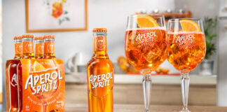 A bottle of Aperol Spritz RTD sits in the middle of a table, with two balloon glasses filled with the cocktail to the bottle's right and a four pack of bottles to the single bottle's left.