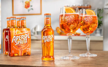 A bottle of Aperol Spritz RTD sits in the middle of a table, with two balloon glasses filled with the cocktail to the bottle's right and a four pack of bottles to the single bottle's left.