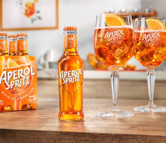 Aperol Spritz comes to the RTD market A bottle of Aperol Spritz RTD sits in the middle of a table, with two balloon glasses filled with the cocktail to the bottle's right and a four pack of bottles to the single bottle's left.