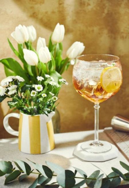 A white and yellow striped vase holds white tulip flowers inside next to a balloon glass with the Marie Brizard Apricot Spritz cocktail inside.