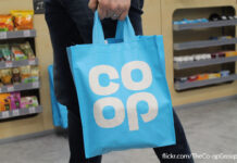 A person carries a blue Co-op Tote bag in a Co-op store with a photograph credit in the bottom right corner.