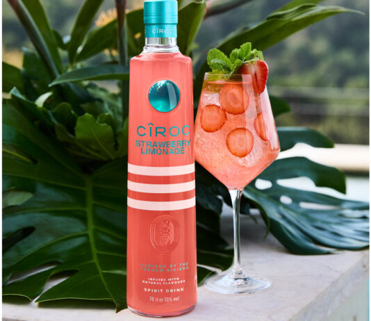 Cîroc introduces limited edition Strawberry Limonade vodka A bottle of Cîroc Strawberry Lemonade stands next to a wine glass with a cocktail inside it made using the vodka with slices of strawberry in the drink as well. These both sit on a stone wall with greenery behind them.