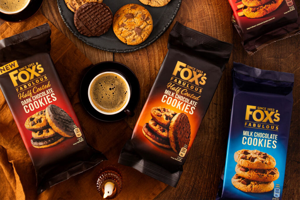 Packets of Fox's Fabulous biscuits sit on a table with biscuits placed onto plates with a mug of coffee between two packs of the biscuits – the biscuit variants include Half Coated Dark Chocolate Cookies, Half Coated Milk Chocolate Cookies, Milk Chocolate Cookies.