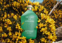 A bottle of Height of Arrows gin lies in a bush of yellow flowers.