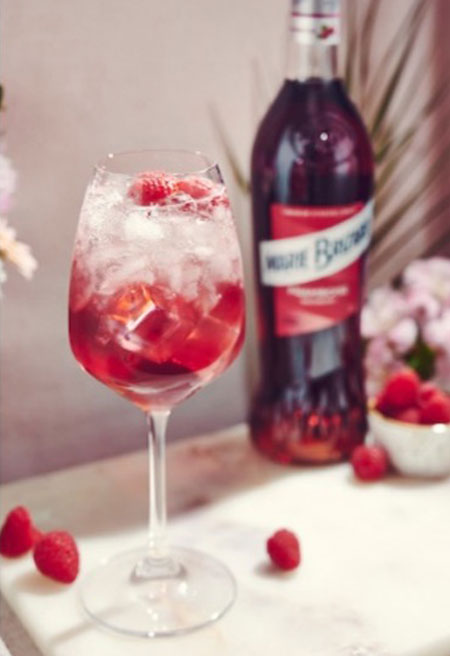 A bottle of Marie Brizard Raspberry Syrup sits next to a balloon glass with Marie Brizard Berry Spritz serve in it. This is served on a white marble table with raspberries on the table and in a small white bowl.