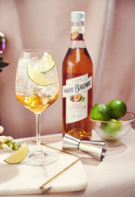 A bottle of Marie Brizard Passionfruit Syrup stands next to a metal jigger measure with a glass bowl of limes next to it. The Marie Brizard Passionfruit Spritz cocktail is served in a balloon glass with a slice of lime next to it on a marble surface.