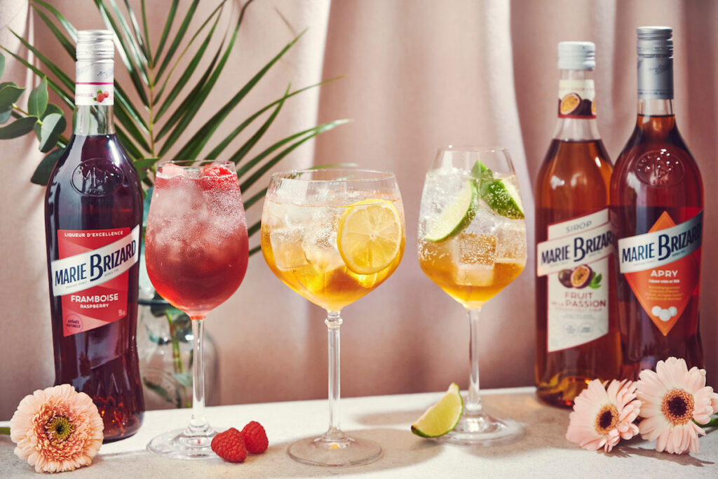 Bottle of Marie Brizar Liqueurs and Sirops stands on a table with cocktails serves in balloon glasses. There are flowers around the table as well as raspberries and a lime slice. The bottles include Marie Brizard Framboise Raspberry Liqueur, Marie Brizard Passion Fruit Sirop and Marie Brizard Apry Liqueur.