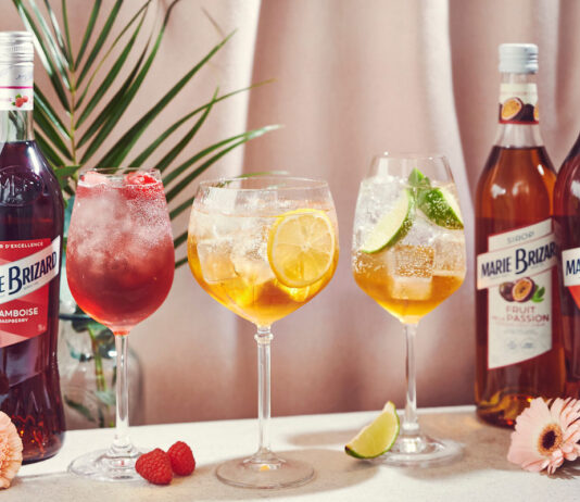 Marie Brizard serves up spring inspiration with Spritz cocktails Bottle of Marie Brizar Liqueurs and Sirops stands on a table with cocktails serves in balloon glasses. There are flowers around the table as well as raspberries and a lime slice. The bottles include Marie Brizard Framboise Raspberry Liqueur, Marie Brizard Passion Fruit Sirop and Marie Brizard Apry Liqueur.