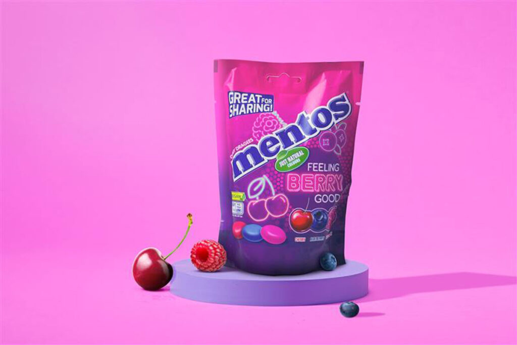 A pack of Mentos Feeling Berry Good sits on a purple coloured podium with pink walls around it. A raspberry also sits on a podium, with a cherry leaning on it and a berry rolling in front of it.